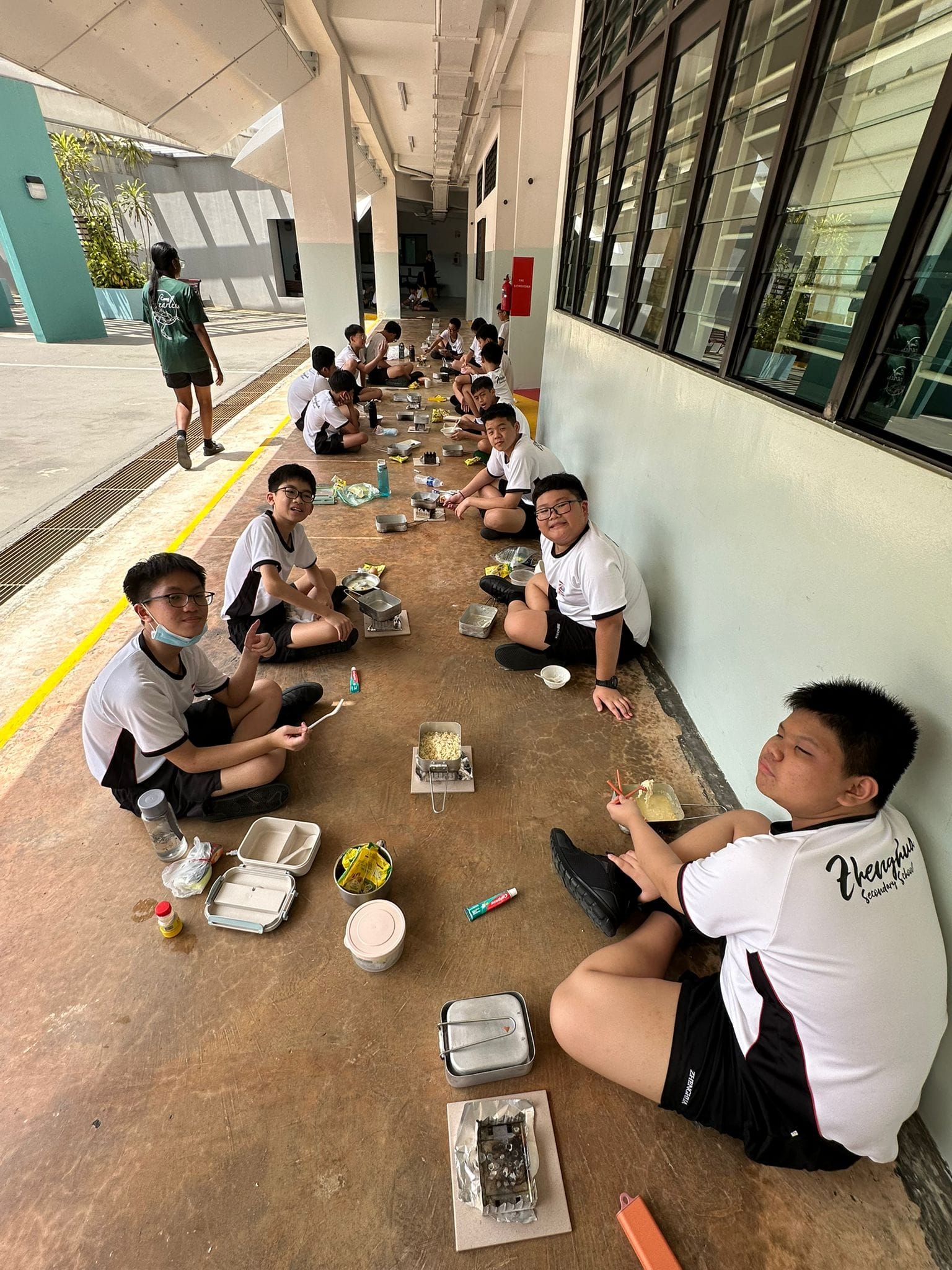 Students in white uniforms sit along a school corridor floor eating meals from metal containers.